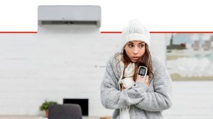 אישה צעירה קופאת מקור בבגדים חמים בבית מחזיקה שלט רחוק עם מזגן ברקע מזגן | צילום רקע: Depositphotos | עיבוד צילום: שולי סונגו ©