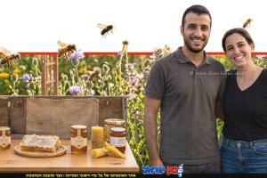 זוג צרכנים עם צנצנת דבש מוזהבת לצד כפפת עץ ייעודית לדבש ותפוחים אדומים על שולחן עץ כפרי | עיבוד: Gemini Google | עיצוב: שולי סונגו©